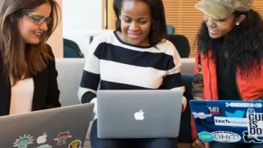 Three women sitting on a sofa working on their laptops.