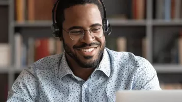 Man wearing headphones, smiling while looking at a laptop in a library.