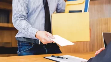 Person handing an envelope to a seated colleague in an office setting.