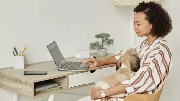 Woman working on laptop with dog in lap at home office.