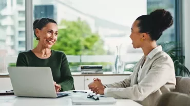 Two women engaged in discussion at a table with a laptop open between them. 