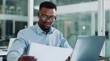 Un homme portant des lunettes et une chemise élégante regarde un document tout en travaillant sur son ordinateur portable.