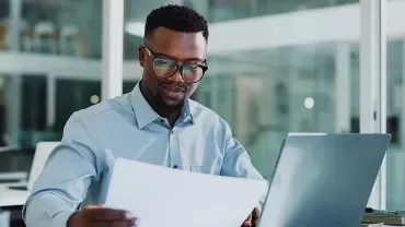 a man with glasses and an elegant shirt looking at a paper and being on his laptop