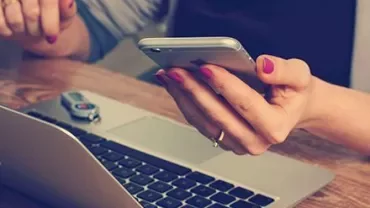 Persoon houdt een smartphone boven een laptop op een houten tafel.