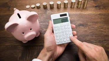 Personne utilisant une calculatrice près d'une tirelire et de piles de pièces.