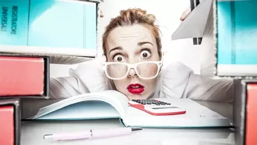 Woman with glasses looks stressed surrounded by binders and an open book.
