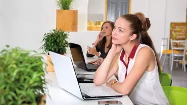 Two women at a desk with laptops, one looking at the screen, the other on a phone call.