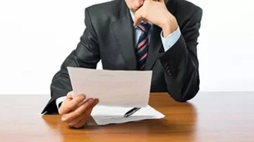 Man in suit reading a document at a wooden desk.