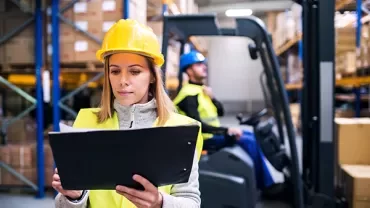 Werknemer in magazijn controleert een clipboard terwijl een ander een vorkheftruck bedient.