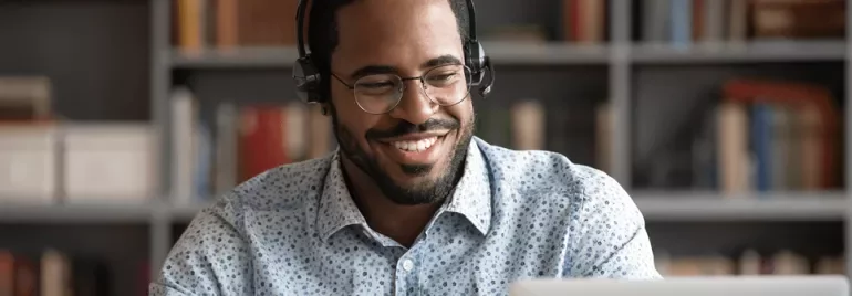 Homme portant des écouteurs, souriant devant un ordinateur portable dans une bibliothèque.