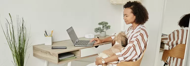 Woman working on laptop with dog in lap at home office.