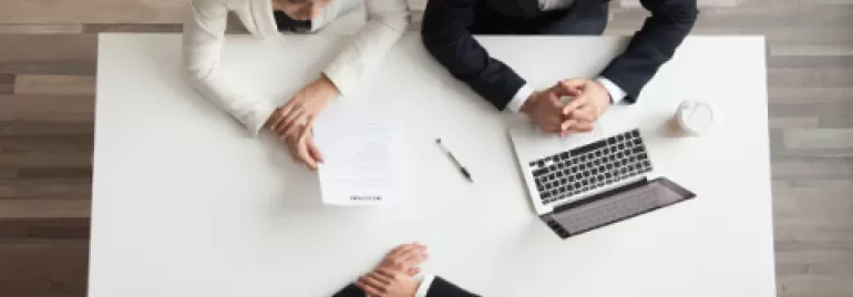 Sollicitatiegesprek met drie mensen aan tafel, documenten en een laptop zichtbaar.