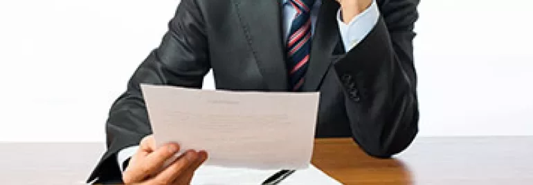 Man in suit reading a document at a wooden desk.