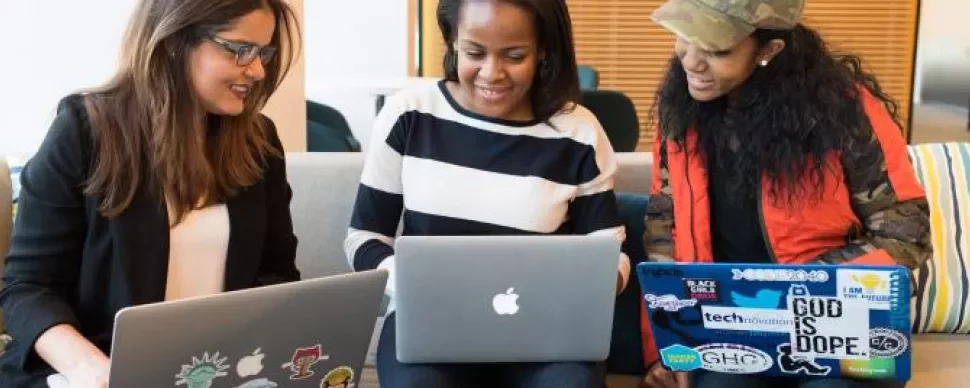 Three women sitting on a sofa working on their laptops.