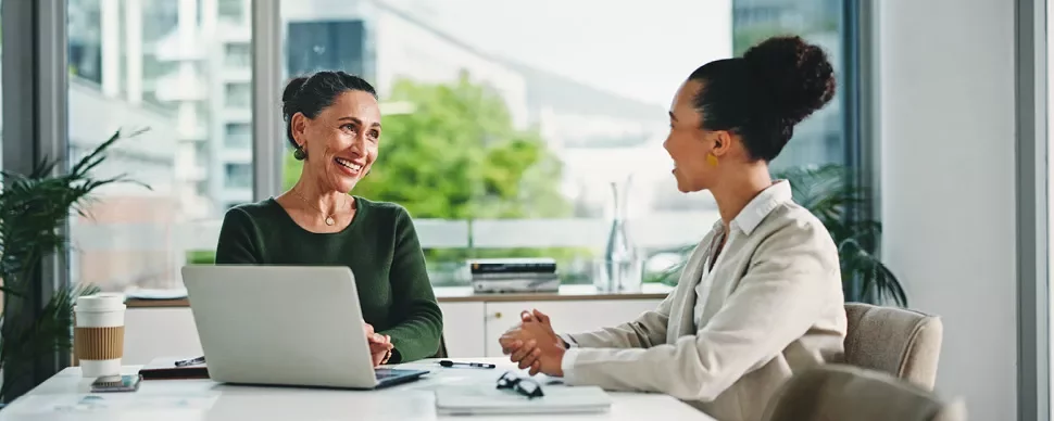 Two women engaged in discussion at a table with a laptop open between them.