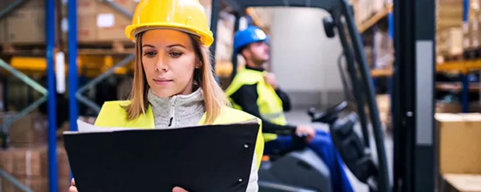 Werknemer in magazijn controleert een clipboard terwijl een ander een vorkheftruck bedient.