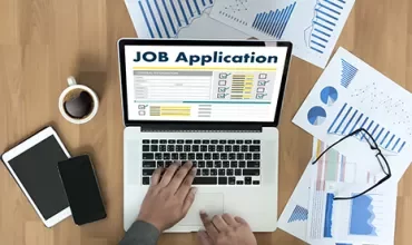 A person typing a job application on a laptop surrounded by charts and papers.
