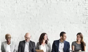 Group of five people sitting against a white brick wall, smiling and holding papers.