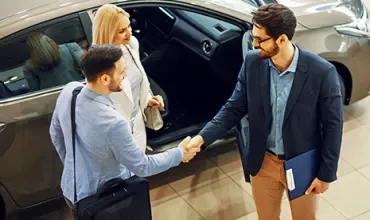 Two men shaking hands next to a car in a dealership.