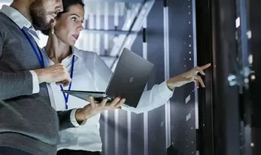 Two people with a laptop in a server room inspecting equipment.