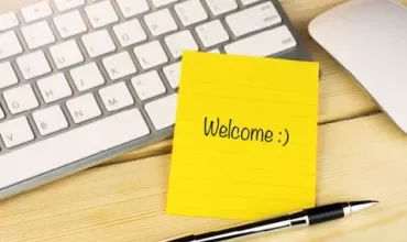 Yellow sticky note with 'Welcome :)' on a desk beside a keyboard and pen.