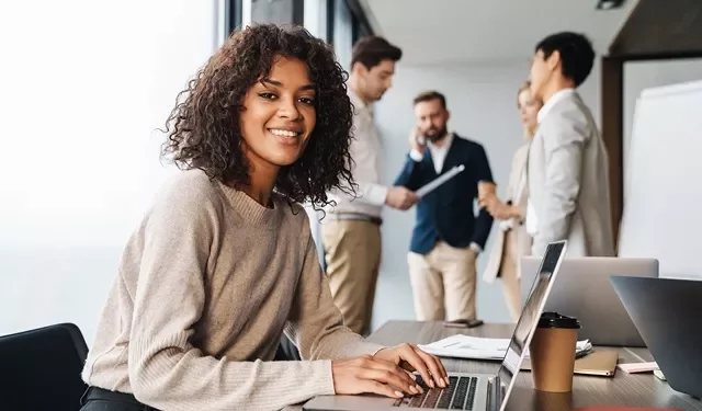 Vrouw glimlacht aan bureau met laptop, collega's op de achtergrond.
