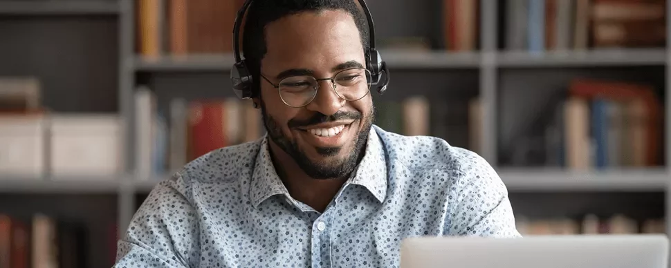 Man wearing headphones, smiling while looking at a laptop in a library.
