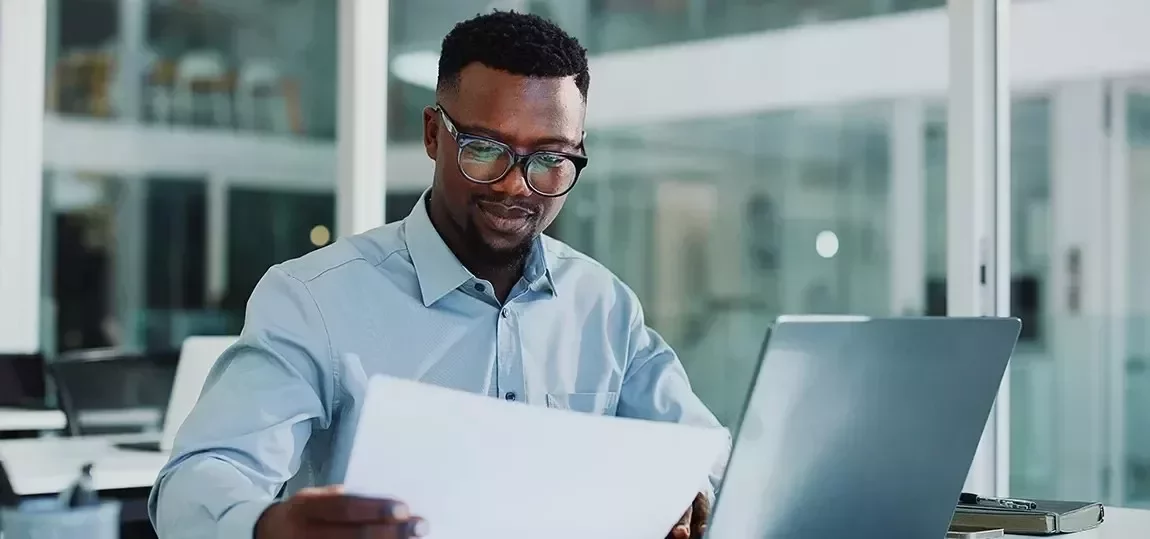 Een man met een bril en een elegant hemd kijkt naar een document terwijl hij op zijn laptop werkt.