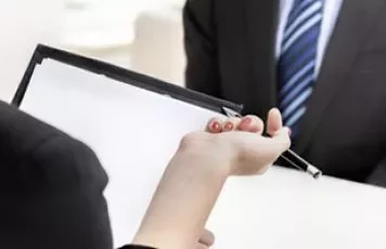 Two people in a meeting, one holding a clipboard with documents.